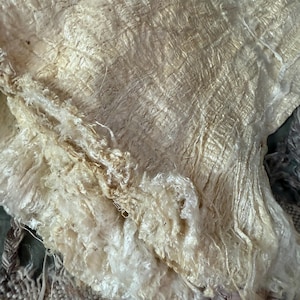 May include: Close-up of a textured, off-white fabric with a layered, organic appearance. The fabric has a woven texture with visible fibers and a slightly frayed edge. The color is a warm, creamy white.