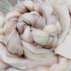 May include: Close-up of a braided bundle of soft, cream-colored wool roving with subtle pink and beige streaks. The wool is arranged in a circular pattern, suggesting a craft project or textile art. A light-colored fabric is in the background.