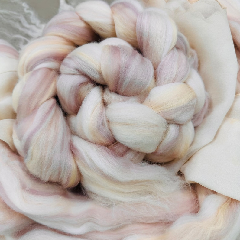 May include: Close-up of a braided bundle of soft, cream-colored wool roving with subtle pink and beige streaks. The wool is arranged in a circular pattern, suggesting a craft project or textile art. A light-colored fabric is in the background.