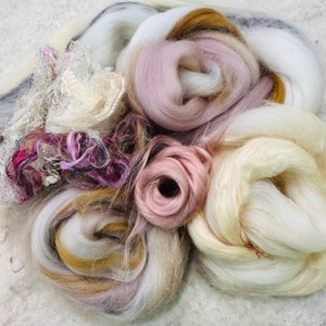 May include: A close-up of a pile of colorful fiber art materials. The fibers are in shades of white, pink, gold, and purple. Some fibers are loosely spun, while others are tightly wound. There are also pieces of lace and ribbon.
