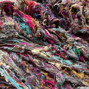 May include: A close-up of a pile of colorful, tangled yarn. The yarn is a mix of blues, pinks, purples, and browns. It appears to be made of recycled materials.