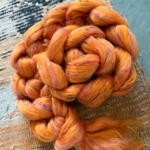 May include: A large, braided skein of vibrant orange yarn with flecks of pink and yellow. The yarn is thick and textured, ideal for knitting or other fiber arts projects. The yarn is displayed on a neutral-toned woven surface.