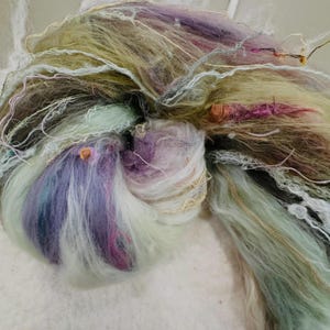 Puede incluir: Primer plano de una madeja de hilo art&iacute;stico en una variedad de colores. El hilo presenta tonos de lavanda, verde y crema, con hebras doradas y rosas. La textura es suave y esponjosa, ideal para proyectos de arte textil.