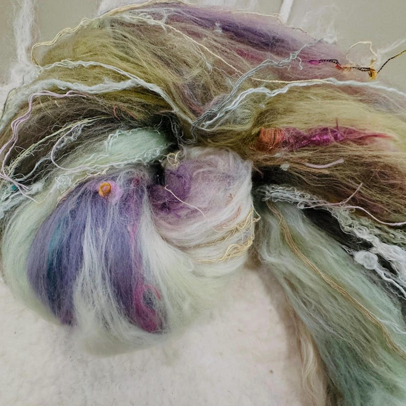 Puede incluir: Primer plano de una madeja de hilo art&iacute;stico en una variedad de colores. El hilo presenta tonos de lavanda, verde y crema, con hebras doradas y rosas. La textura es suave y esponjosa, ideal para proyectos de arte textil.