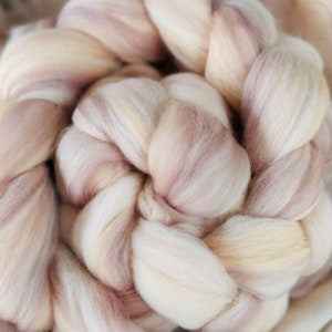 May include: Close-up of a braided bundle of soft, undyed wool roving. The wool fibers are a blend of creamy white, pale pink, and light beige, creating a natural, textured appearance. Ideal for fiber art projects.