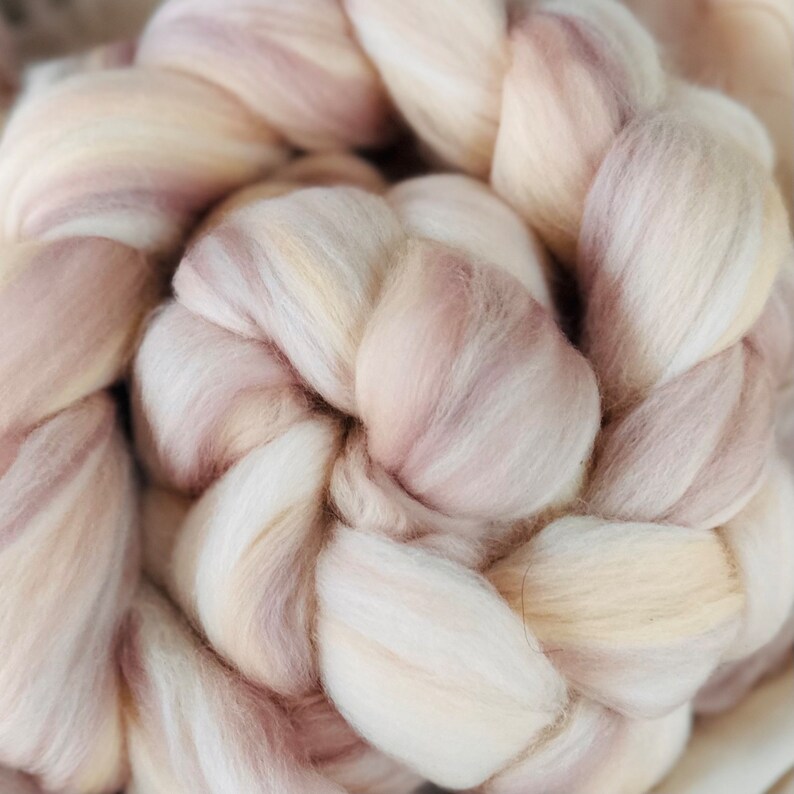 May include: Close-up of a braided bundle of soft, undyed wool roving. The wool fibers are a blend of creamy white, pale pink, and light beige, creating a natural, textured appearance. Ideal for fiber art projects.