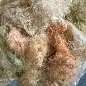 May include: A collection of loose, fibrous material with a tangled, web-like texture. The fibers display a mix of pale cream, soft beige, and muted peach tones, creating a layered, airy appearance with fine, delicate strands throughout the mass.