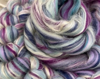 Può includere: Primo piano di un gomitolo di filato colorato con sfumature di viola, blu, bianco e crema. Il filato è disposto in un motivo a spirale, creando una texture visivamente accattivante.