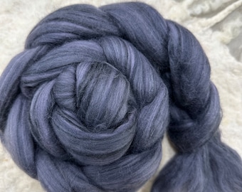 Può includere: Primo piano di un gomitolo di roving di lana grigio scuro, strettamente arrotolato e intrecciato. La lana presenta sottili variazioni di colore, con tonalità più chiare e scure che creano un aspetto strutturato. Lo sfondo è una superficie chiara e strutturata.