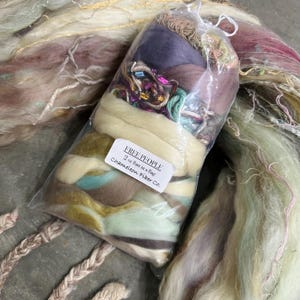 May include: A clear plastic bag filled with a variety of colorful fibers, including purple, green, and brown. The bag is labeled "FREE PEOPLE 2 oz Batt in a Bag Chameleon Fiber Co."