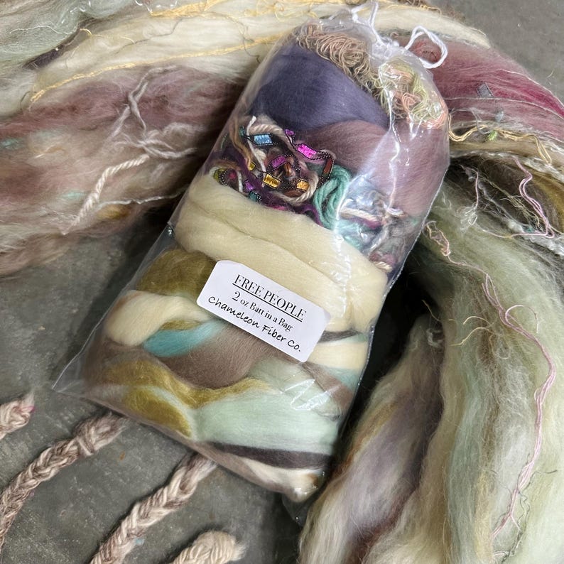 May include: A clear plastic bag filled with a variety of colorful fibers, including purple, green, and brown. The bag is labeled "FREE PEOPLE 2 oz Batt in a Bag Chameleon Fiber Co."