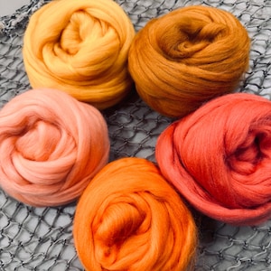May include: Four balls of wool yarn in shades of orange, peach, and red. The yarn is arranged in a circle on a gray mesh surface.