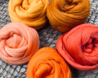 May include: Four balls of wool yarn in shades of orange, peach, and red. The yarn is arranged in a circle on a grey mesh surface.