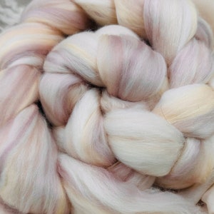 May include: Close-up of a skein of hand-dyed wool roving. The wool is a blend of cream, pale pink, and light brown, creating a soft, textured appearance. The fibers are arranged in a braided pattern, ideal for spinning or felting.