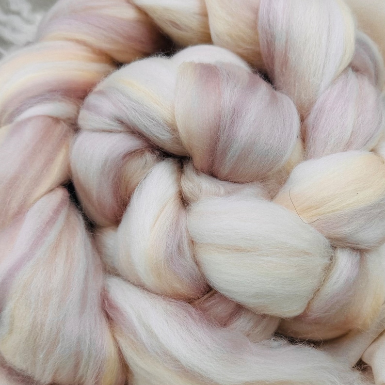 May include: Close-up of a skein of hand-dyed wool roving. The wool is a blend of cream, pale pink, and light brown, creating a soft, textured appearance. The fibers are arranged in a braided pattern, ideal for spinning or felting.