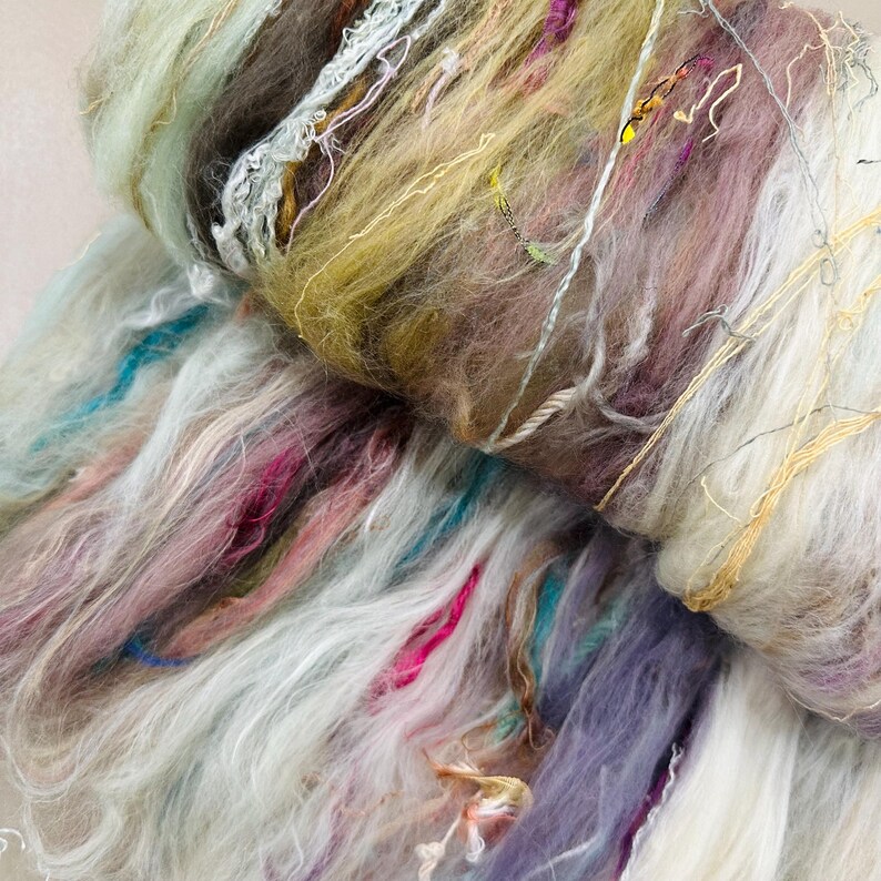 Puede incluir: Primer plano de un ovillo de lana colorido y texturizado. La lana presenta una mezcla de colores, incluyendo blanco, turquesa, rosa, morado y amarillo. Los hilos de diferentes grosores y texturas crean una apariencia suave y en capas, ideal para proyectos de arte textil.