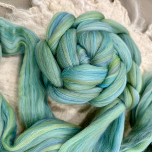 May include: A close-up of a skein of hand-dyed wool roving in shades of teal, blue, and green. The wool is arranged in a circular pattern, with strands extending outwards. The background is a neutral, off-white fabric.