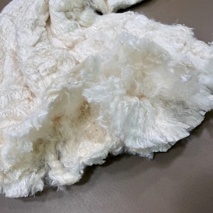 May include: A cream-colored, textured knit garment featuring a thick, shaggy, and fibrous surface. The material displays a dense, layered appearance with long, soft-looking strands that create a voluminous and highly tactile finish throughout.