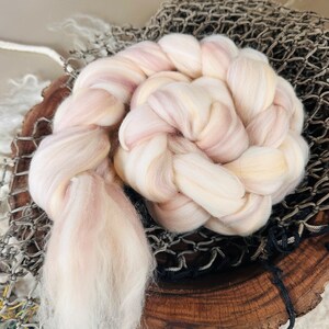 May include: A braided skein of pastel-colored wool roving, featuring soft pink and cream hues. The fiber is arranged in a circular pattern, with a portion unraveled. The wool is displayed on a wooden surface with a fishing net.