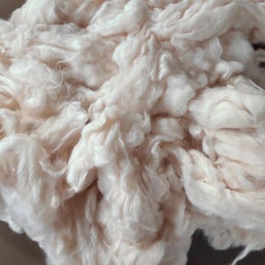 May include: A dense, fluffy pile of raw, unspun off-white cotton fibers. The material features a soft, fibrous texture with visible natural variations in the off-white color, showing the loose, tangled structure of the raw plant material.