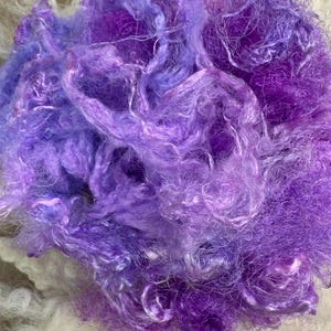 May include: A loose, unspun mass of fiber featuring a vibrant blend of purple and violet hues. The material has a soft, wispy, and slightly shiny texture with varying densities of tangled strands, creating a cloud-like appearance throughout.