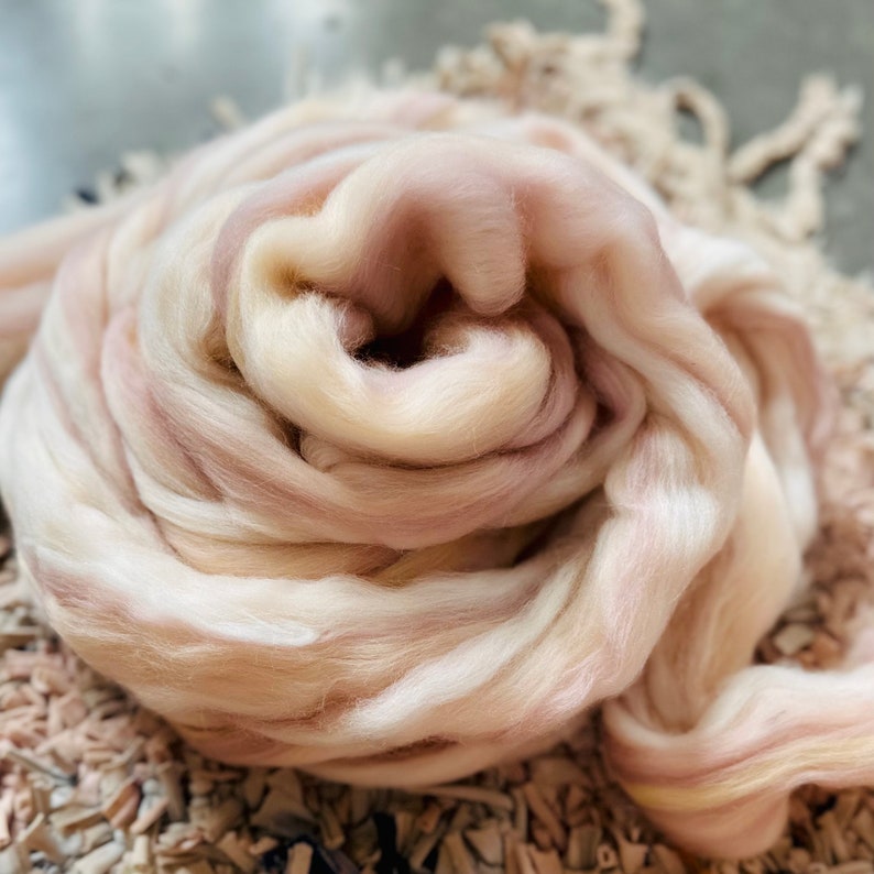 May include: A swirl of soft, undyed wool roving in shades of cream, pale pink, and light brown. The fibers are loosely spun, creating a textured appearance. This wool is ideal for various fiber art projects, including spinning, felting, and weaving.