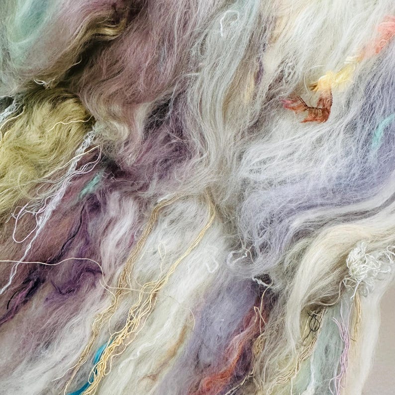 Puede incluir: Primer plano de un mont&oacute;n de fibra colorida. Las fibras son una mezcla de blanco, lavanda, morado y verde p&aacute;lido, con hebras doradas y azules. La textura parece suave y esponjosa, adecuada para manualidades.