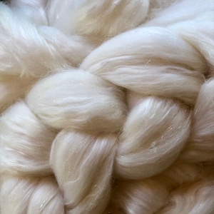 May include: A close-up of a white, fluffy, and sparkly fiber. The fiber is arranged in a braided pattern, creating a soft and luxurious texture.