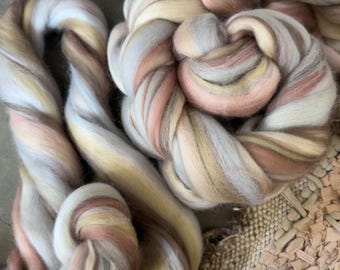May include: A close-up of a bundle of soft, multi-colored yarn. The yarn is a mix of white, light pink, light brown, and beige. The yarn is arranged in a loose, flowing knot.
