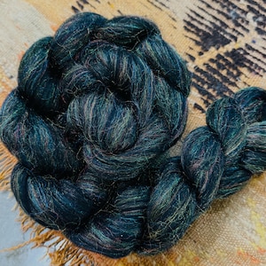 May include: A dark teal-colored skein of yarn, tightly wound and braided. The yarn has subtle iridescent highlights of pink, gold, and green. The fiber appears soft and luxurious, suitable for knitting or other fiber arts projects.