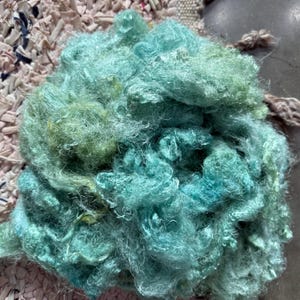 May include: A close-up of a pile of teal and green fiber. The fiber has a soft, fluffy texture and appears to be dyed in shades of turquoise and seafoam green. The fibers are loosely arranged, creating a textured, organic appearance.