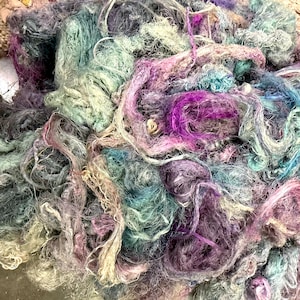 May include: A close-up of a pile of colorful yarn in shades of purple, blue, green, and cream. The yarn appears to be a mix of textures and thicknesses, creating a visually interesting and tactile display. The yarn is likely used for knitting or other fiber crafts.