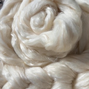 May include: A coiled bundle of off-white, lustrous fiber with a smooth, silky texture. The long, continuous strands are arranged in a circular, layered pattern, reflecting light with a subtle sheen across the soft, fibrous surface of the material.