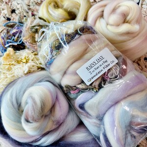 May include: A clear plastic bag filled with a 2 oz batt of fiber in a variety of colors, including white, purple, blue, and pink. The bag is labeled "Rainy Day 2 oz Batt in a Bag Chameleon Fiber Co."