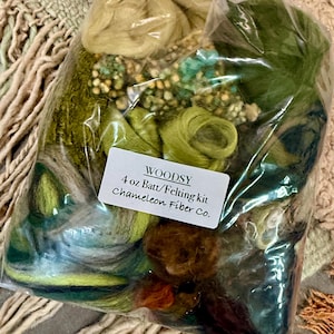 May include: A clear plastic bag containing assorted green, brown, and cream-colored wool fibers for felting. A white label on the front reads: WOODSY 4 oz Batt/Felting kit Chameleon Fiber Co. The fibers have varied textures, including loose tufts and coils.