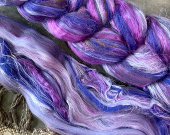 Può includere: Primo piano di fibra tinta a mano nei toni del viola, blu e bianco. La fibra è disposta in una treccia e in fili sciolti, mostrando le variazioni di colore e le trame. La fibra viene probabilmente utilizzata per filare, tessere o altre arti tessili.