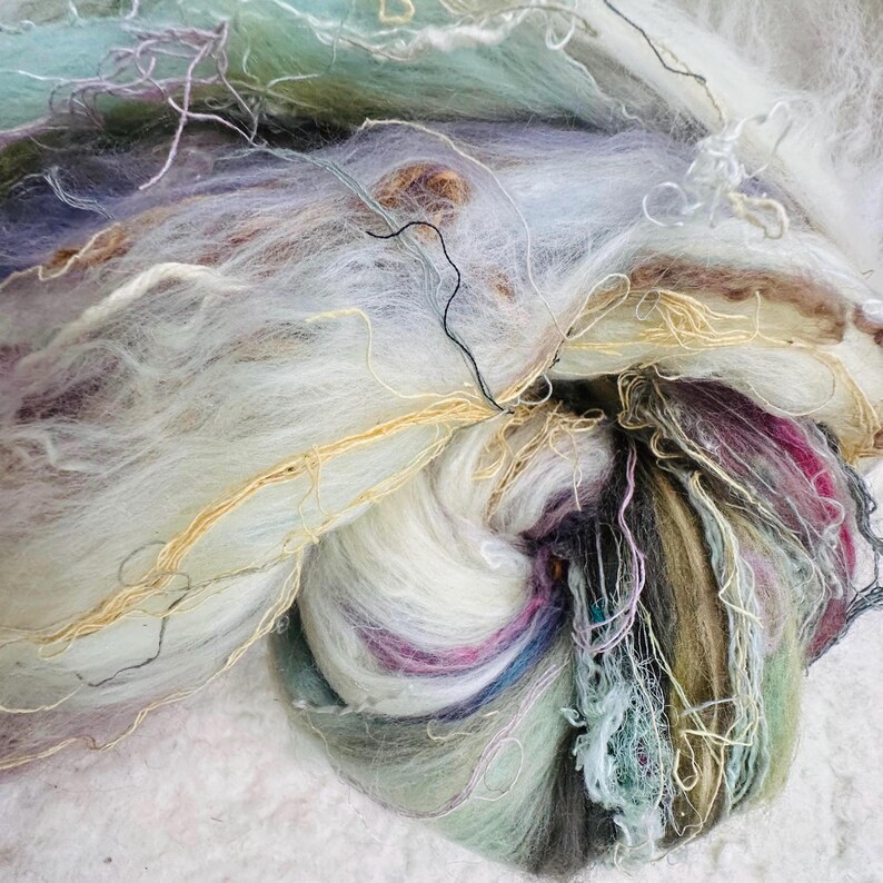 Puede incluir: Primer plano de un ovillo de hilo art&iacute;stico con una mezcla de colores. El hilo presenta una mezcla de blanco, crema y tonos pastel, incluyendo azul claro, morado y verde. Finos hilos dorados y negros est&aacute;n entrelazados.