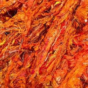 May include: Close-up of a pile of textured, orange-colored yarn. The yarn has a variety of textures, including thick, thin, and knotted strands. The color palette ranges from light orange to deep reddish-orange, with hints of pink and yellow.