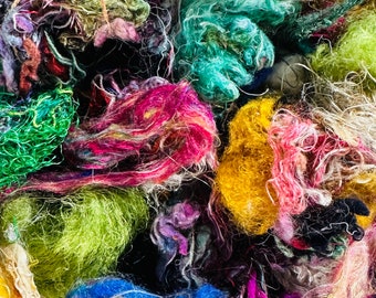 May include: A close-up of a pile of colorful yarn scraps in various shades of green, blue, yellow, pink, and red. The yarn is soft and fluffy, and it appears to be made from natural fibers.