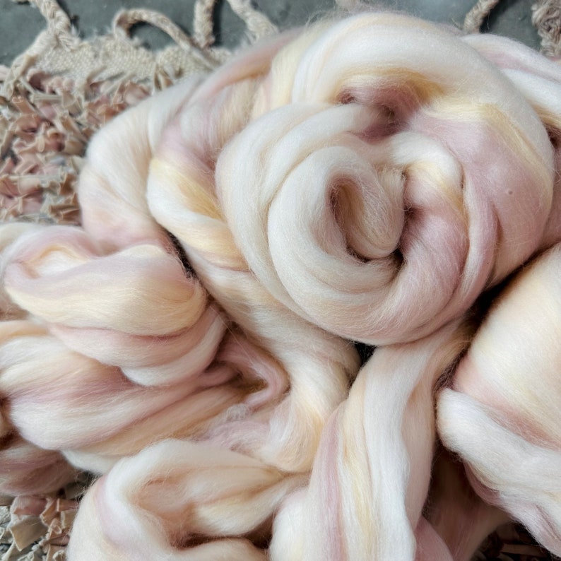 May include: Close-up of a pile of soft, undyed wool roving. The wool is a blend of creamy white, pale yellow, and light pink hues. The fibers are loosely spun, creating a textured appearance. Ideal for fiber art projects.