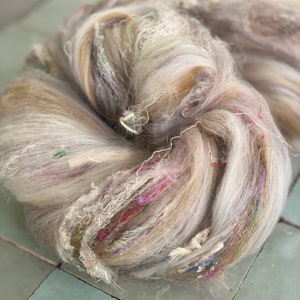 May include: A large, loosely wound skein of yarn in shades of gray, beige, and cream. The yarn has a soft, fluffy texture with strands of colorful threads, including pink, green, and blue. The yarn is on a light blue tiled surface.