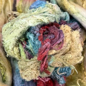 May include: A bundle of hand-dyed silk ribbon yarn featuring a textured, frayed appearance. The fibers display a variegated color palette including shades of sage green, deep burgundy, dusty blue, and golden tan, woven into a loose, tangled cluster.