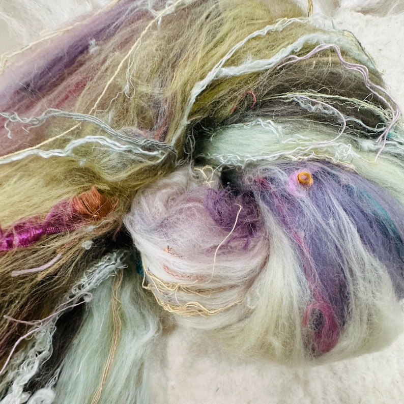 Puede incluir: Primer plano de un ovillo de hilo colorido en tonos lavanda, verde y crema. El hilo tiene un aspecto suave y texturizado con hebras de diferentes grosores y texturas. Algunas hebras tienen un brillo met&aacute;lico.