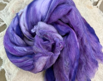 May include: A close-up of a bundle of purple and white fiber. The fiber is soft and fluffy, with a mix of light and dark purple hues. The fiber is sitting on a white, textured surface.