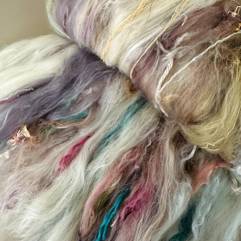 Puede incluir: Primer plano de un ovillo de lana colorida. La lana presenta una mezcla de colores, incluyendo blanco, morado, rosa, turquesa y dorado. La textura parece suave y esponjosa, con hebras de diferente grosor. Ideal para proyectos de arte textil.