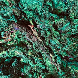 May include: A close-up of a pile of teal, green, and purple silk fibers. The fibers are tangled and interwoven, creating a textured and abstract pattern.