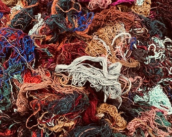 May include: A close-up of a pile of colorful yarn scraps in various shades of red, brown, blue, green, and white. The yarn is tangled and knotted, creating a textured and abstract pattern.