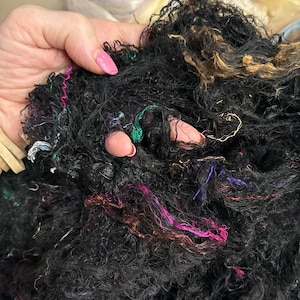 May include: Close-up of a handful of black fiber with colorful strands of pink, green, and gold. The fiber appears to be a blend of materials, possibly for textile or craft projects. The texture is dense and slightly coarse.