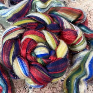 May include: Close-up of colorful hand-spun yarn. The yarn is a mix of red, yellow, blue, and green, with strands of silver glitter. The yarn is arranged in a circular pattern, with some strands extending outwards. The yarn is likely used for knitting or other fiber arts.