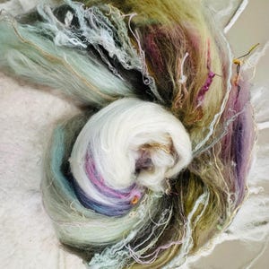Puede incluir: Primer plano de una madeja de hilo te&ntilde;ido a mano en una variedad de colores. El hilo es una mezcla de blanco, azul claro, verde, morado y dorado. El hilo es suave y esponjoso, y parece estar hecho de una fibra natural.